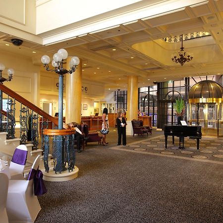 Stamford Grand Adelaide Hotel Glenelg Beltér fotó The photo shows a luxurious interior of a hotel or a restaurant lobby. There is an elegant staircase with ornate railings leading to an upper level. The space features high ceilings with decorative chandeliers and ample natural light coming through l