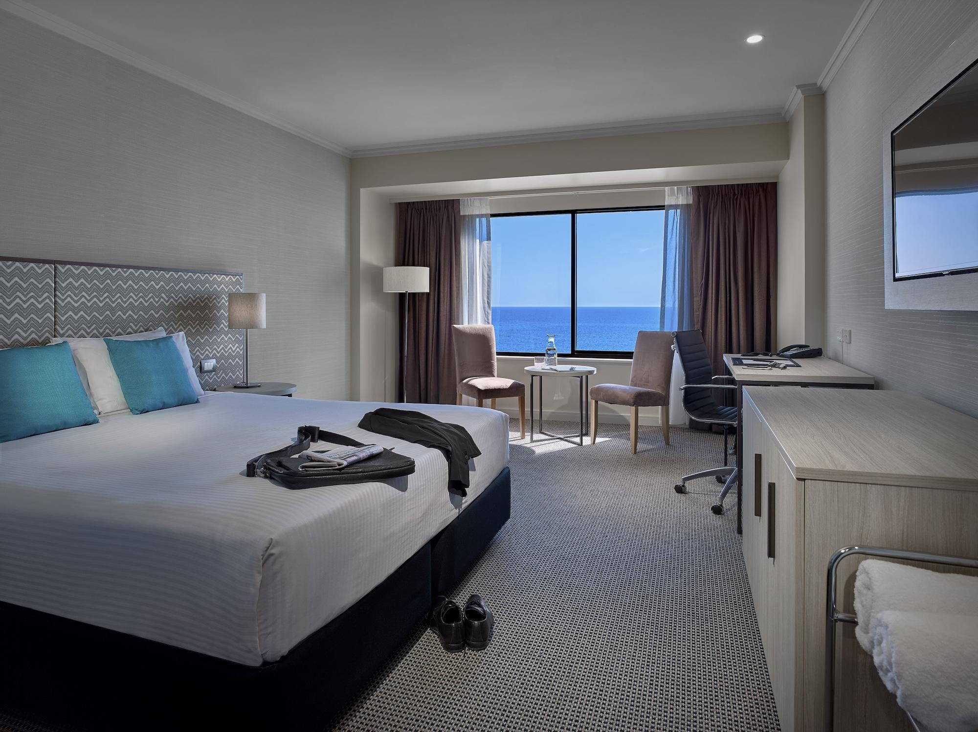 Stamford Grand Adelaide Hotel Glenelg Kültér fotó The photo shows a modern hotel room featuring a queen-sized bed with white linens and two aqua blue decorative pillows. There are bedside lamps and a large window with a view of the ocean. The room also has a small seating area with two chairs and a 