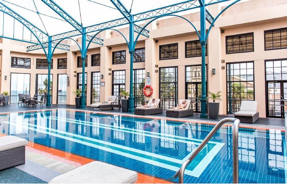 Stamford Grand Adelaide Hotel Glenelg Kültér fotó The photo shows an indoor swimming pool area characterized by a modern and elegant design. The pool features blue tiles with two white stripes running along its length. Surrounding the pool, there are several lounge chairs for relaxation. The walls h
