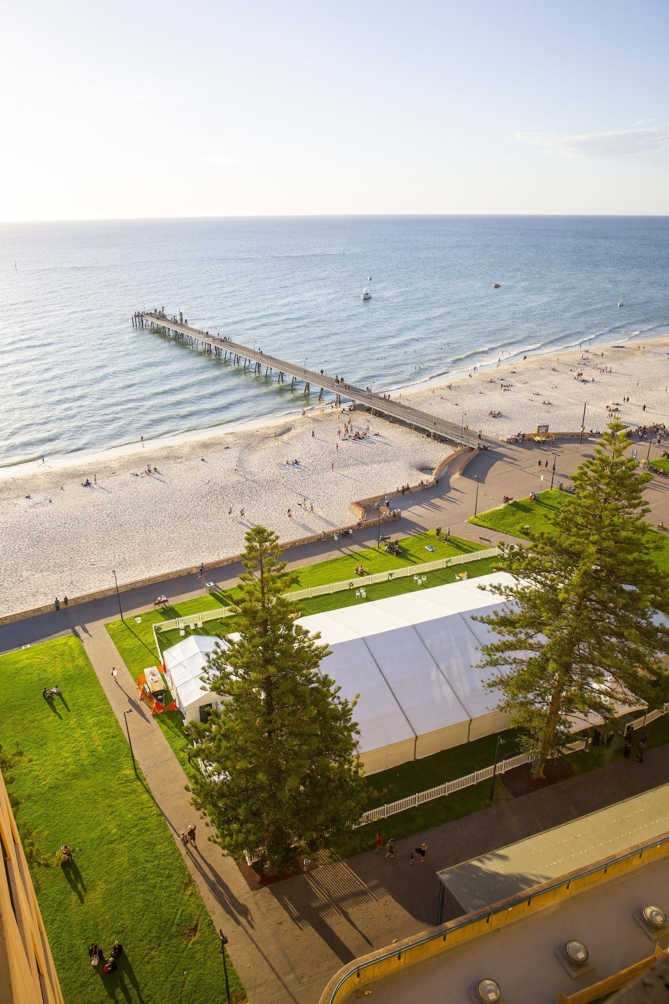 Stamford Grand Adelaide Hotel Glenelg Kültér fotó The photo shows a scenic view of a beach with a long pier extending into the water. The beach is sandy, and there are people enjoying the sun and walking along the shore. In the foreground, there is a large white tent set up on a grassy area, likely 