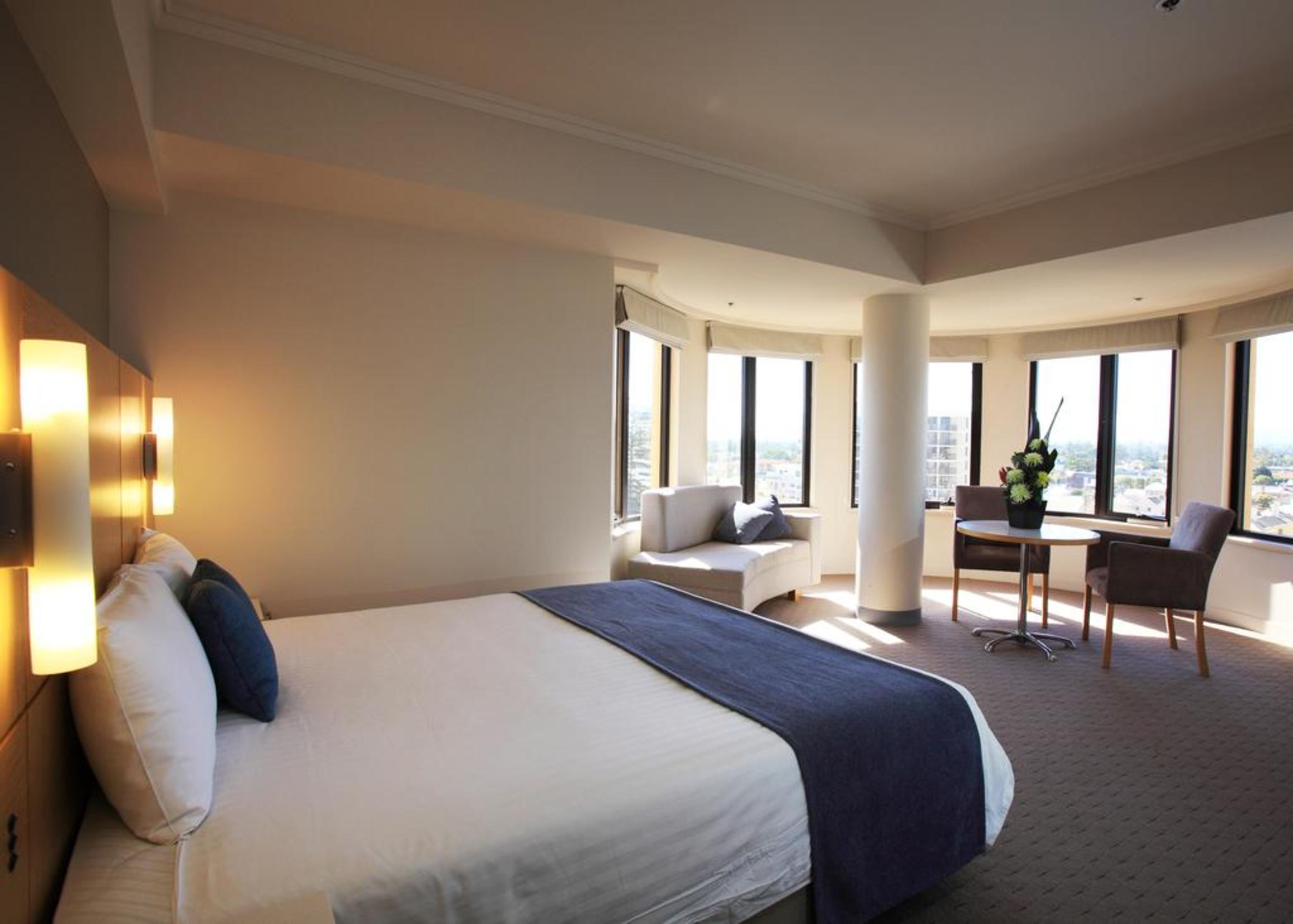 Stamford Grand Adelaide Hotel Glenelg Kültér fotó The photo shows a hotel room with a modern and spacious design. There is a large bed with white linens and a dark blue throw blanket. The room features multiple windows that allow plenty of natural light, and there is a small seating area near the wi