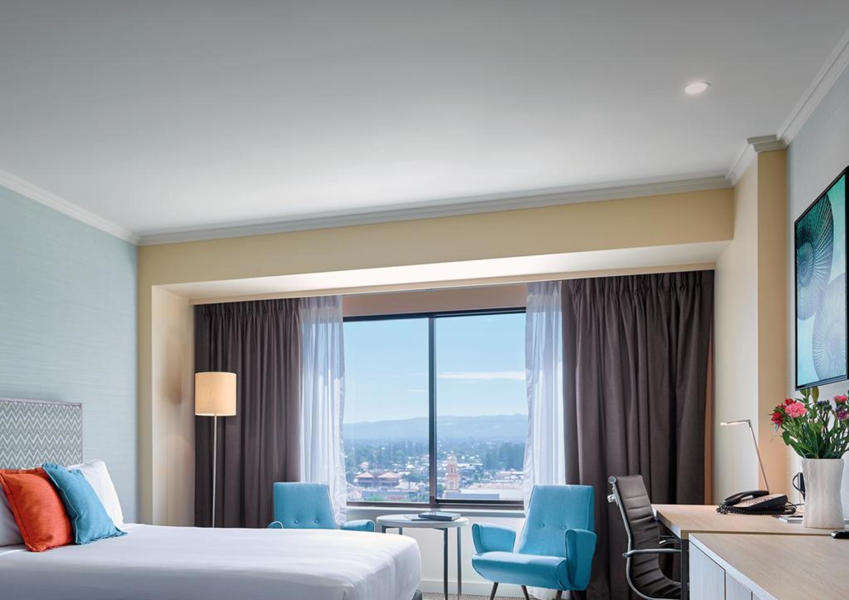 Stamford Grand Adelaide Hotel Glenelg Kültér fotó The photo shows a modern hotel room featuring a queen-sized bed with white linens and colorful decorative pillows. There are two blue chairs beside a small table positioned near a large window that provides a view of the outside, with sheer and dark 