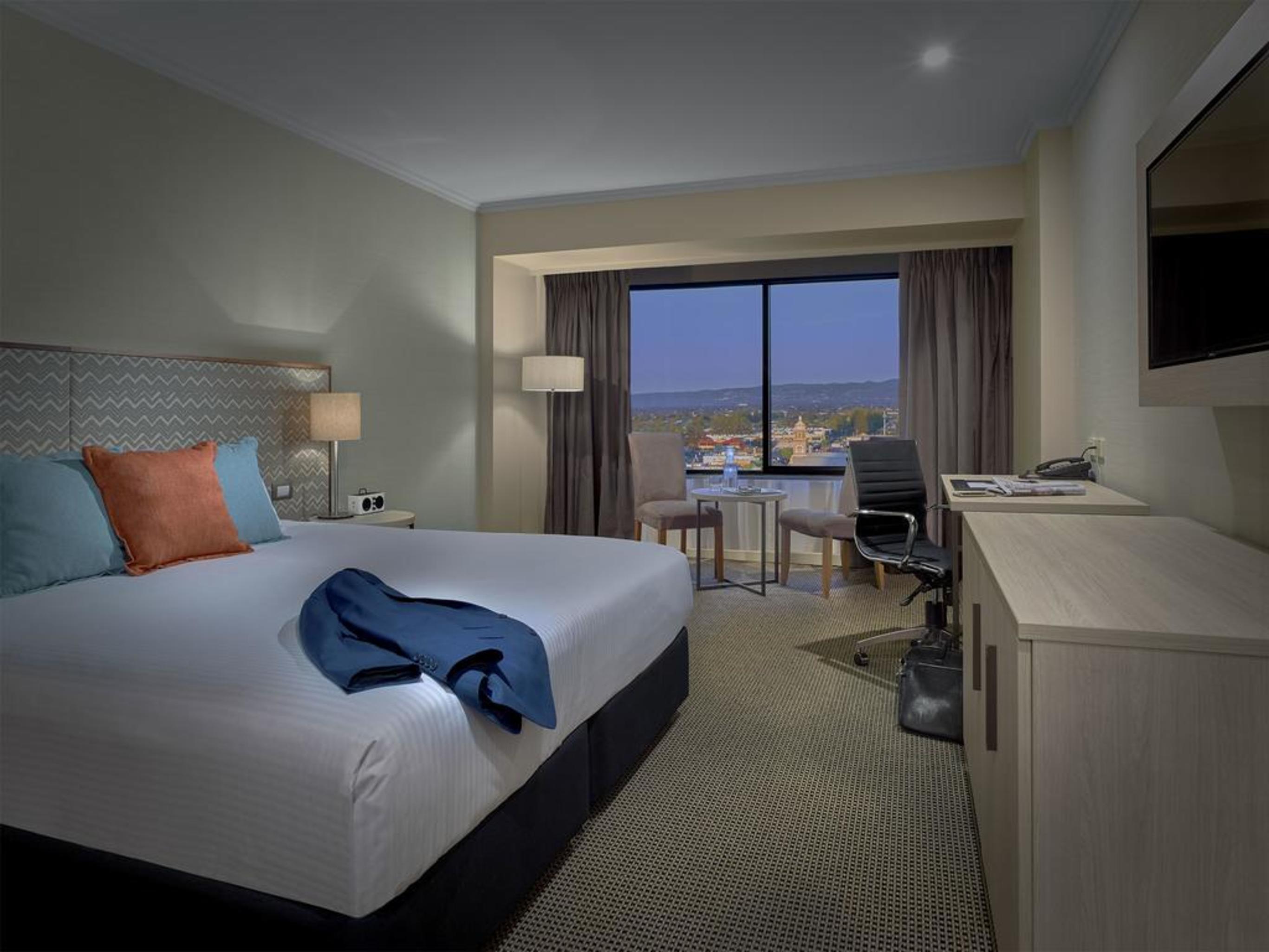 Stamford Grand Adelaide Hotel Glenelg Kültér fotó The photo shows a modern hotel room. It features a large double bed with white bedding and decorative pillows in orange and blue. There's a nightstand with lamps on either side of the bed. A large window overlooks a view outside, and there's a desk a