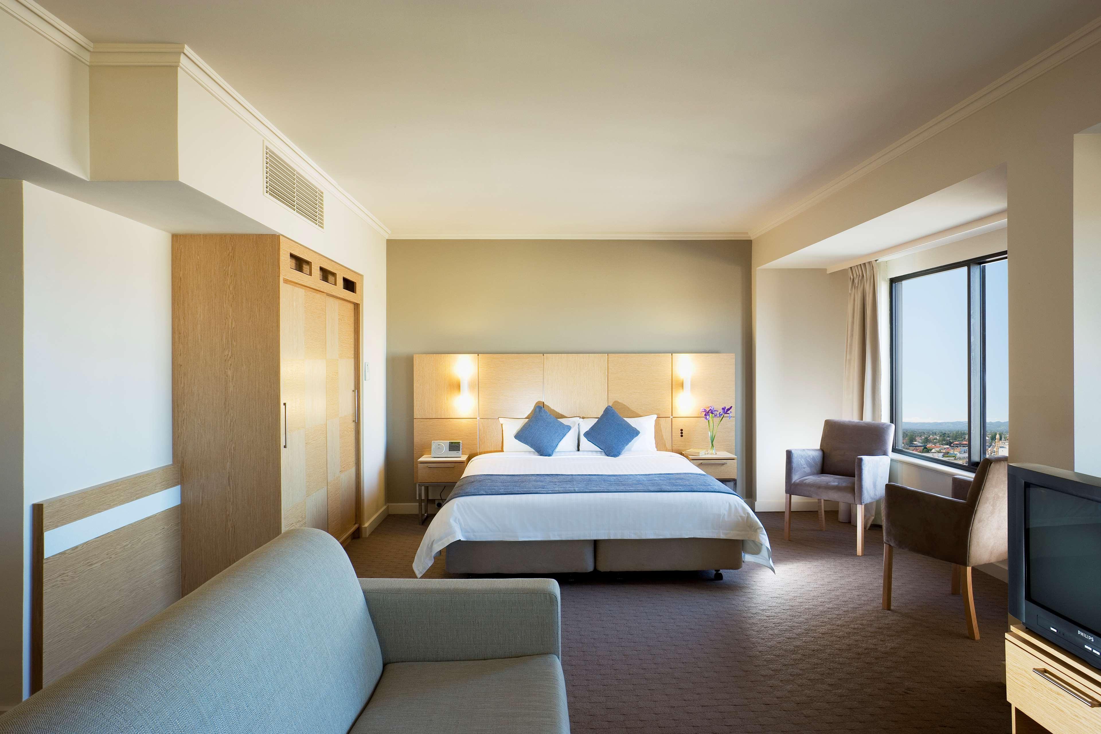 Stamford Grand Adelaide Hotel Glenelg Szoba fotó The photo shows a modern hotel room featuring a large bed with two blue decorative pillows. There are bedside lamps on both sides of the headboard. To the left, there's a wooden wardrobe and a small table with a vase and a few amenities. On the right