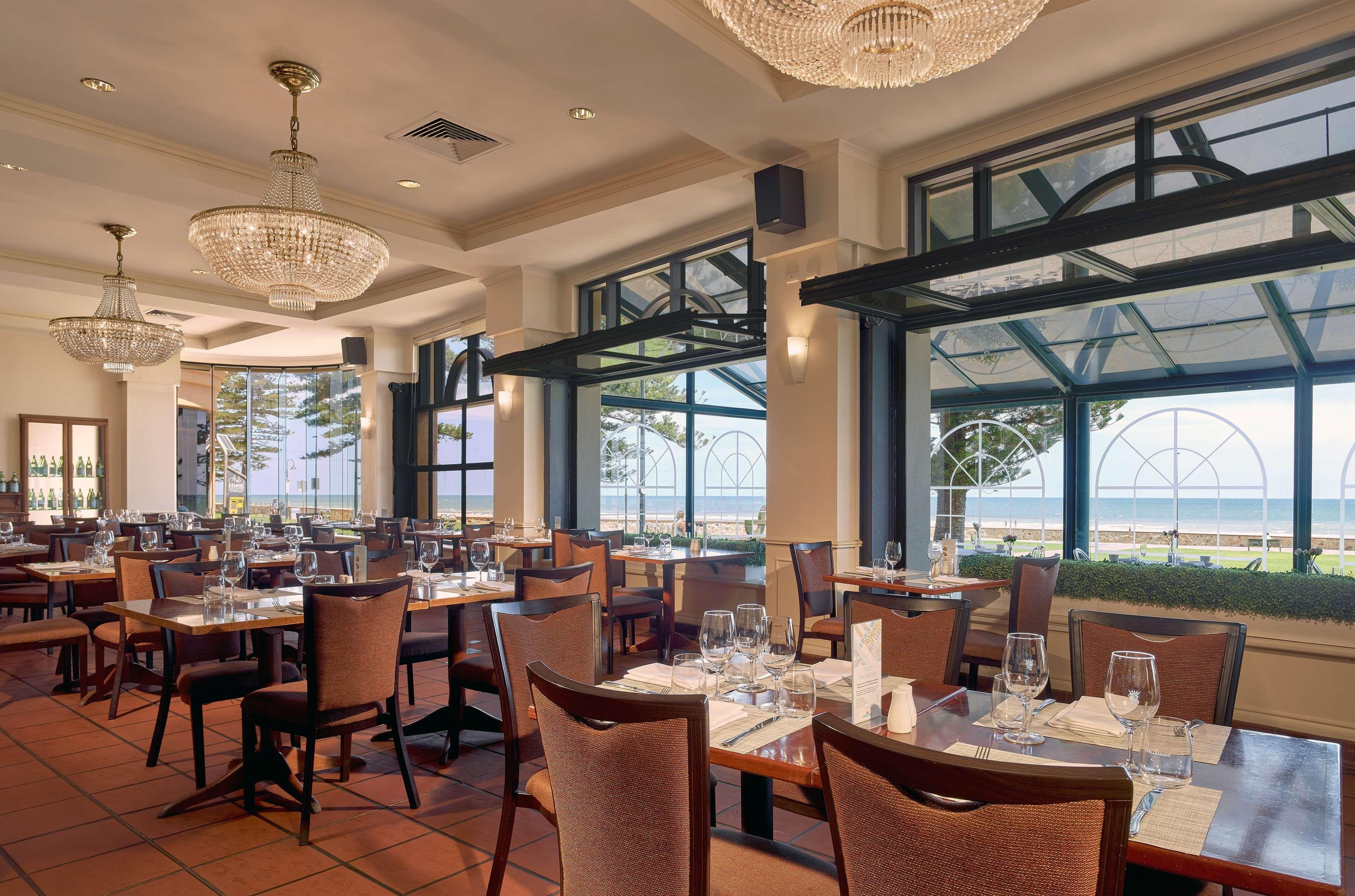 Stamford Grand Adelaide Hotel Glenelg Kültér fotó The photo shows the interior of a restaurant. It features several wooden tables set with glasses and utensils, suggesting it's ready for diners. The decor includes elegant chandeliers hanging from the ceiling, adding a touch of sophistication. Large 