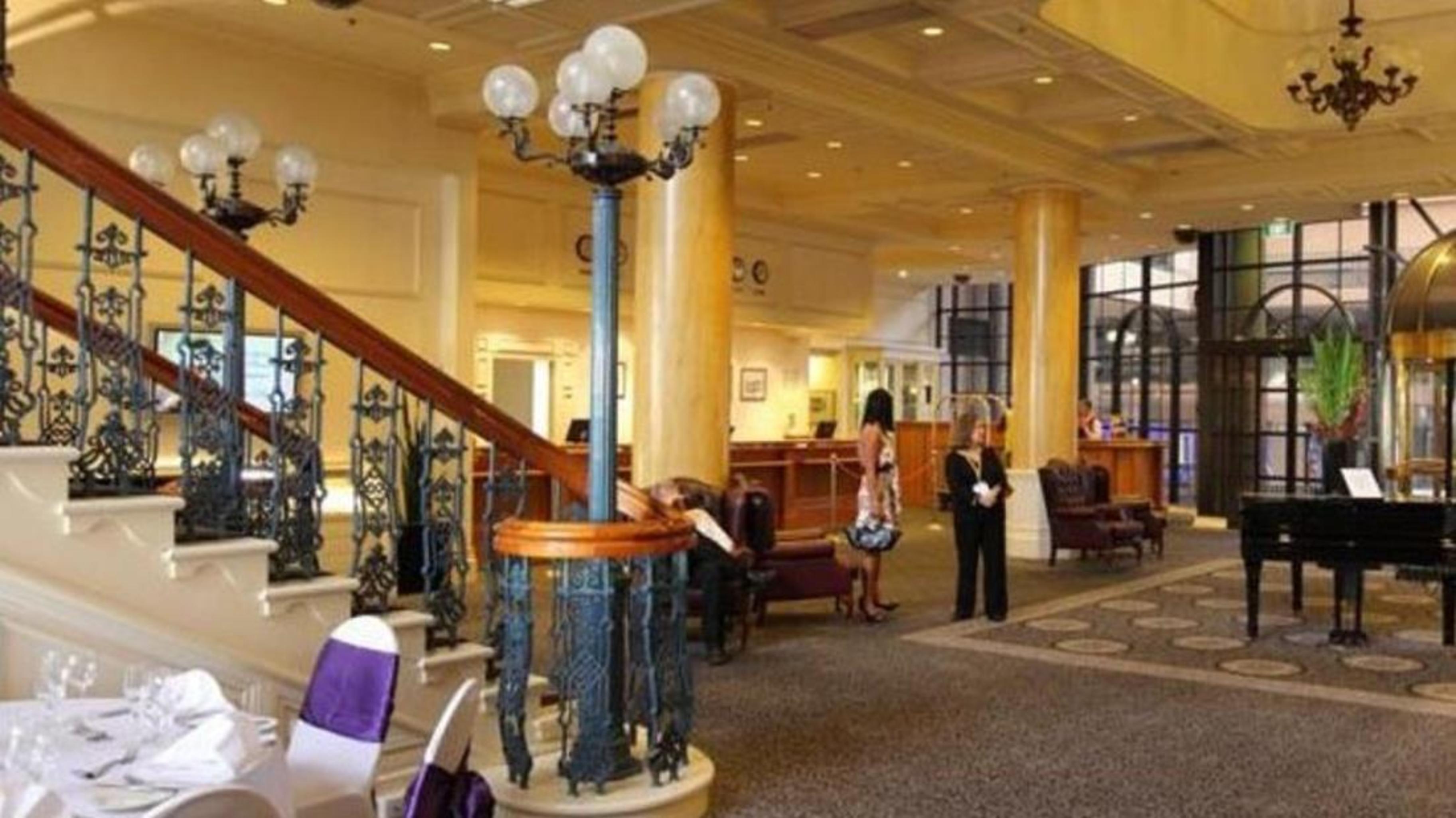 Stamford Grand Adelaide Hotel Glenelg Kültér fotó The photo shows the interior of a hotel lobby. It features a spacious area with a high ceiling and elegant decor. There is a staircase with a decorative railing leading to an upper level. In the foreground, some guests and staff are present, and ther