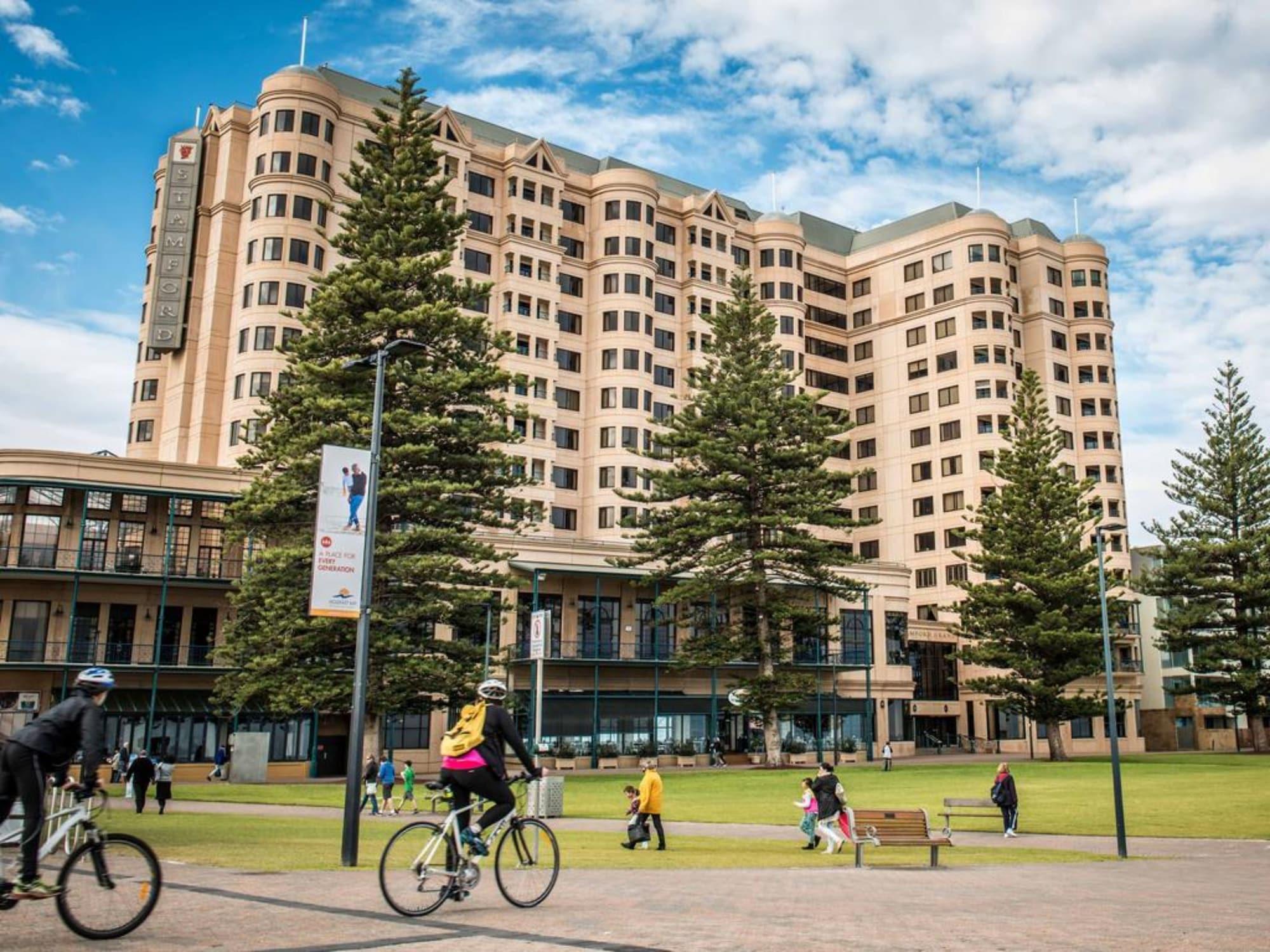 Stamford Grand Adelaide Hotel Glenelg Kültér fotó The photo depicts a modern building with a large facade, likely a hotel or commercial establishment, situated near a spacious green area. Surrounding the building are several tall trees, providing shade and enhancing the aesthetic appeal. In the fore