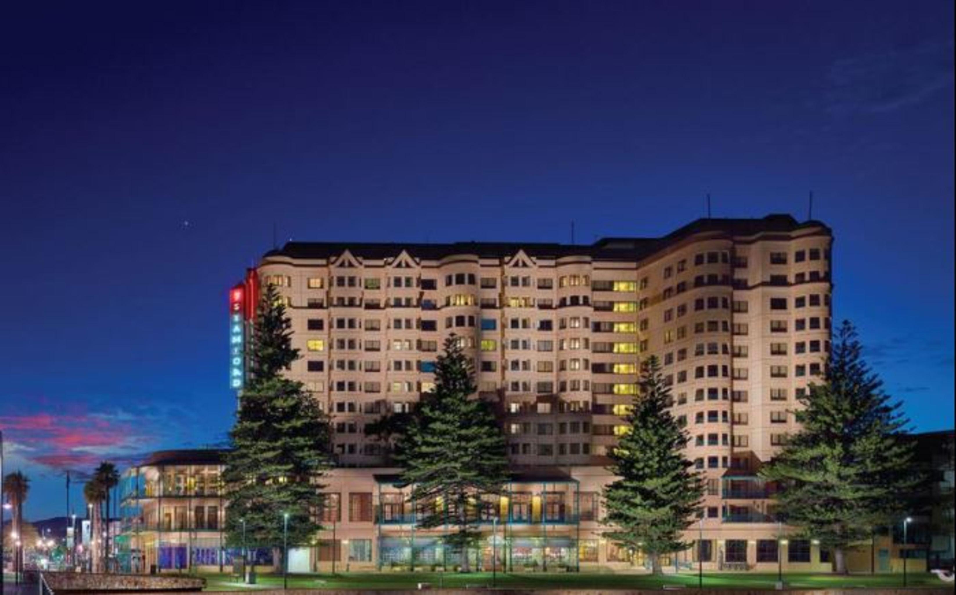 Stamford Grand Adelaide Hotel Glenelg Kültér fotó The photo displays a multi-story hotel building at dusk, illuminated by various lights. The architecture features a mix of modern and traditional design elements, with numerous windows showcasing a warm glow. Surrounding the hotel are several tall tr