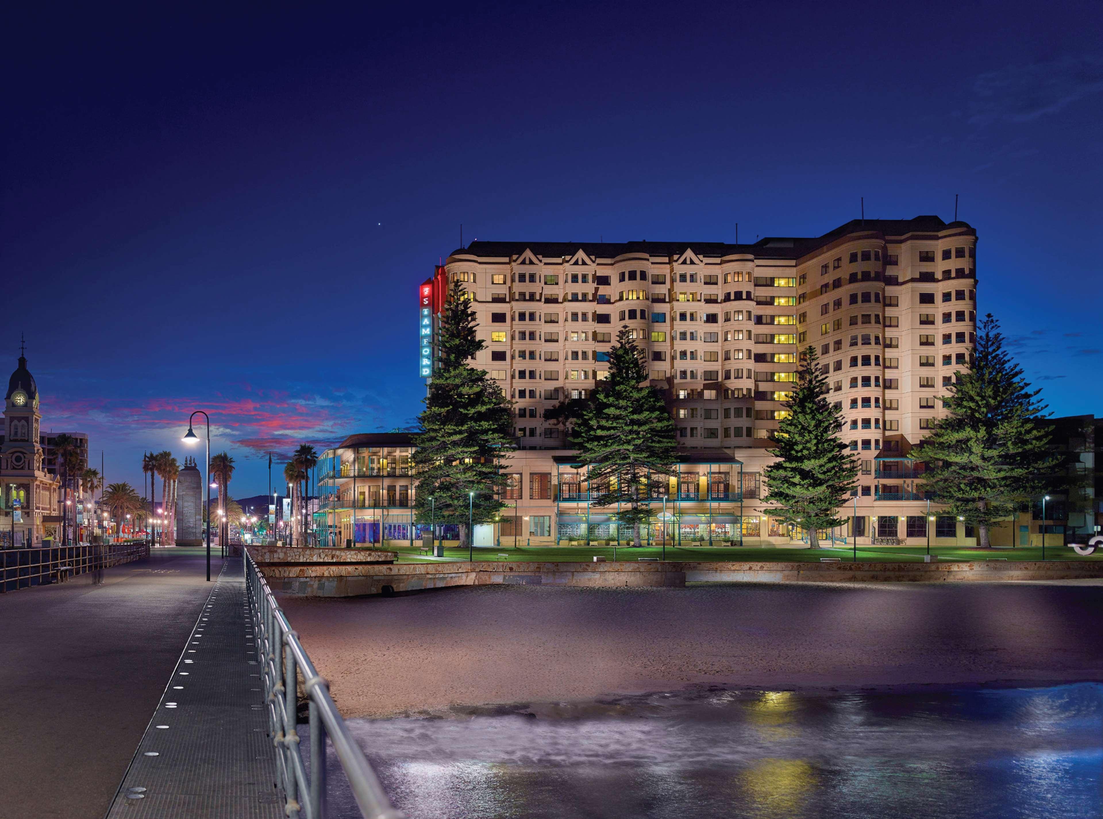 Stamford Grand Adelaide Hotel Glenelg Kültér fotó The photo shows a waterfront scene at dusk, featuring a large building lit up against a darkening sky. The architecture appears modern and prominent, with several windows illuminated. In the foreground, there is a walkway or pier with fencing along t