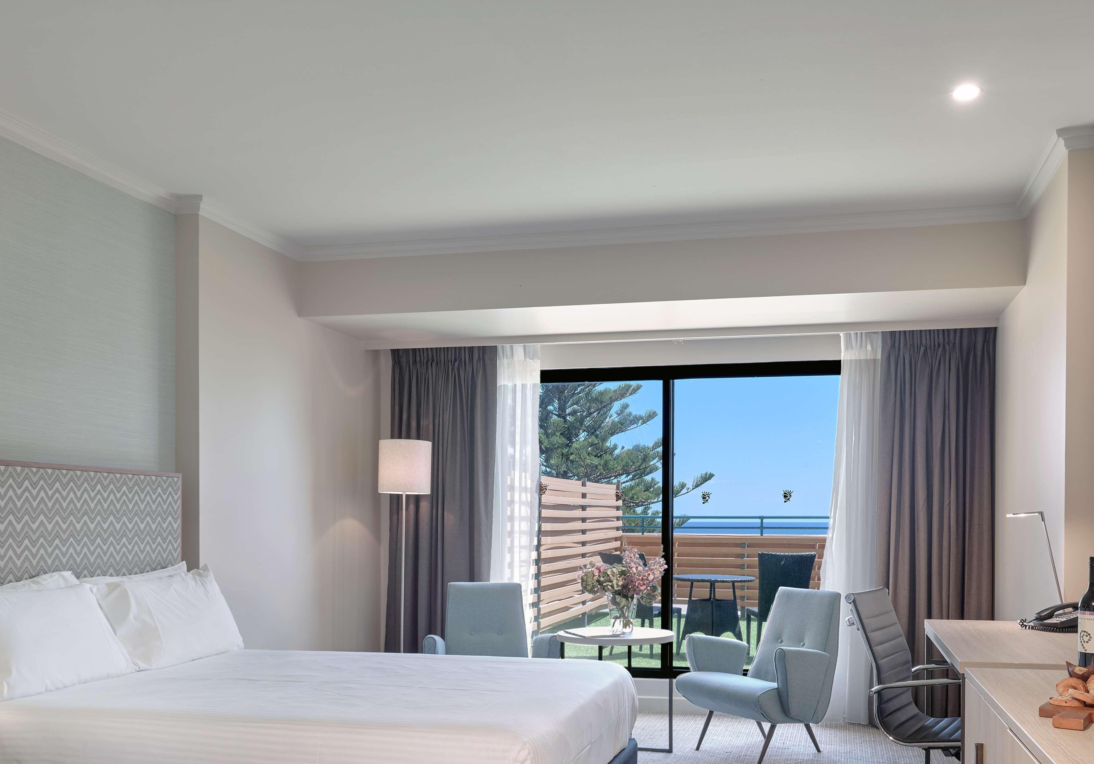 Stamford Grand Adelaide Hotel Glenelg Kültér fotó The photo shows a modern hotel room featuring a large bed with white bedding. There are two armchairs positioned near a window that offers a view of an outdoor area, possibly a balcony or patio with greenery. The room has light-colored walls and curt