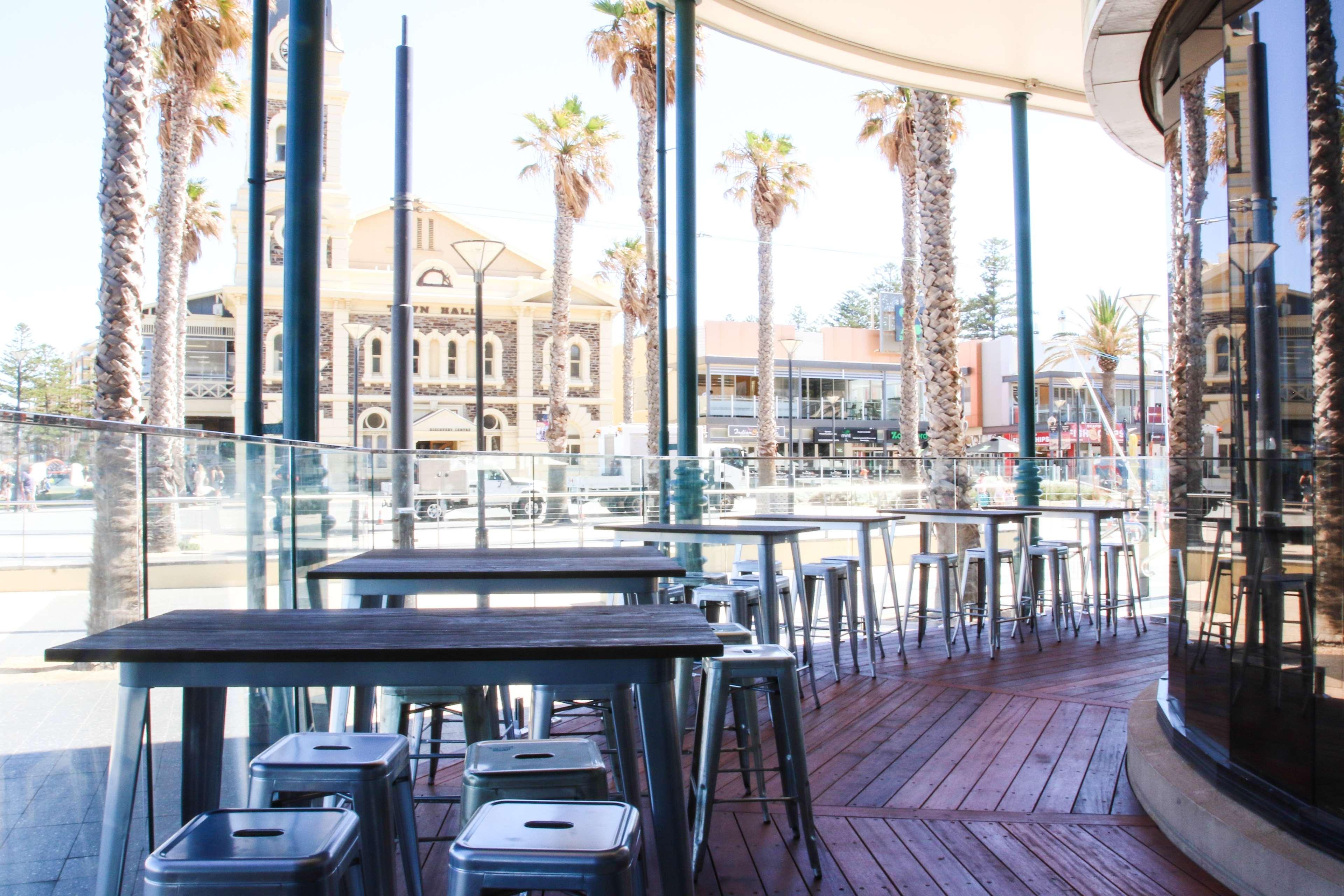 Stamford Grand Adelaide Hotel Glenelg Kültér fotó The photo shows an outdoor seating area of a café or restaurant. It features metal tables and stools arranged on a wooden deck. The setting has large glass panels, providing an open and airy atmosphere. In the background, palm trees and a building wi