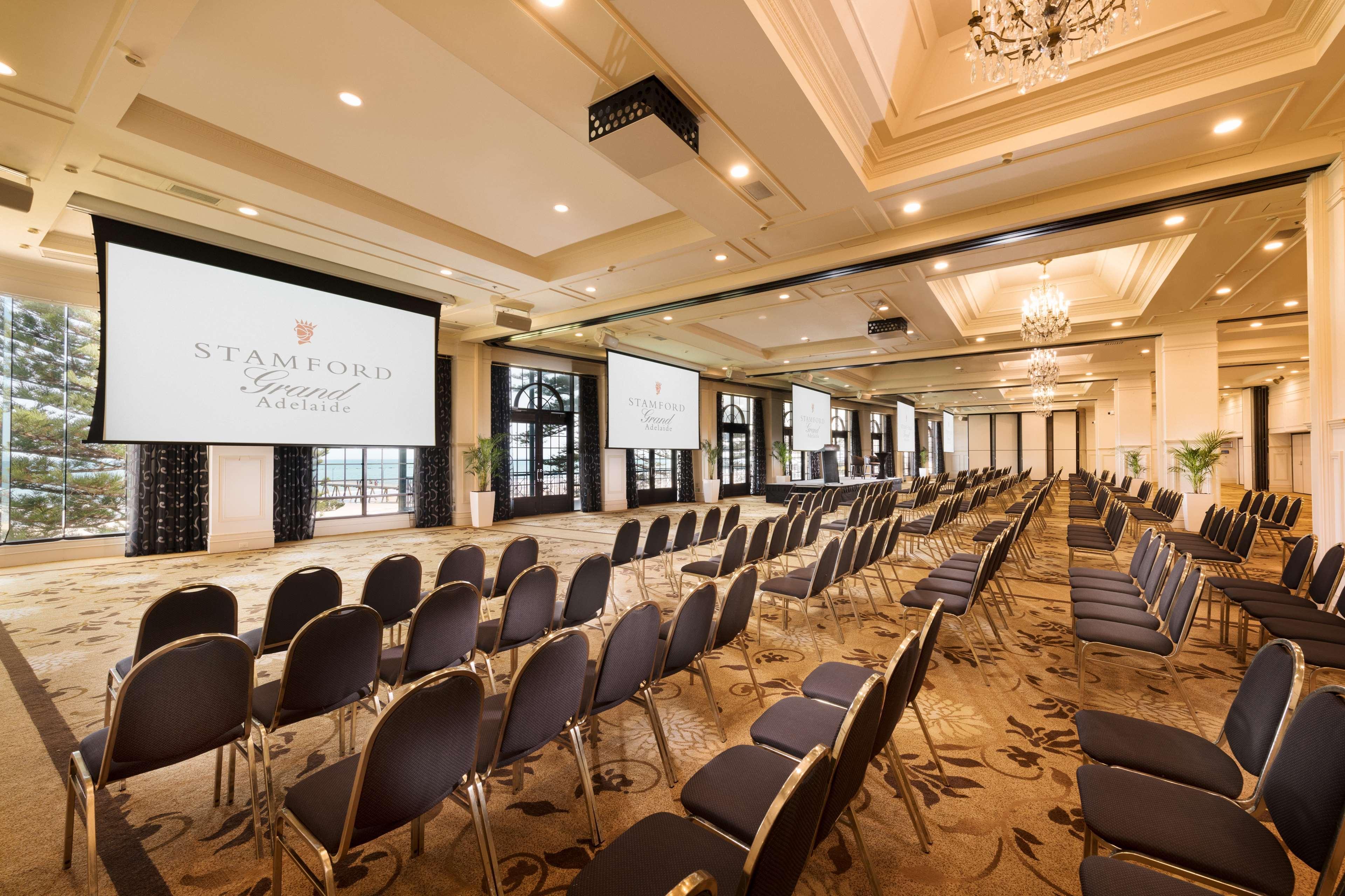 Stamford Grand Adelaide Hotel Glenelg Kültér fotó The photo shows a spacious event or conference room. There are rows of dark, upholstered chairs arranged facing two large screens. The room features elegant decor with soft lighting, a chandelier, and windows that allow natural light to filter in. Th