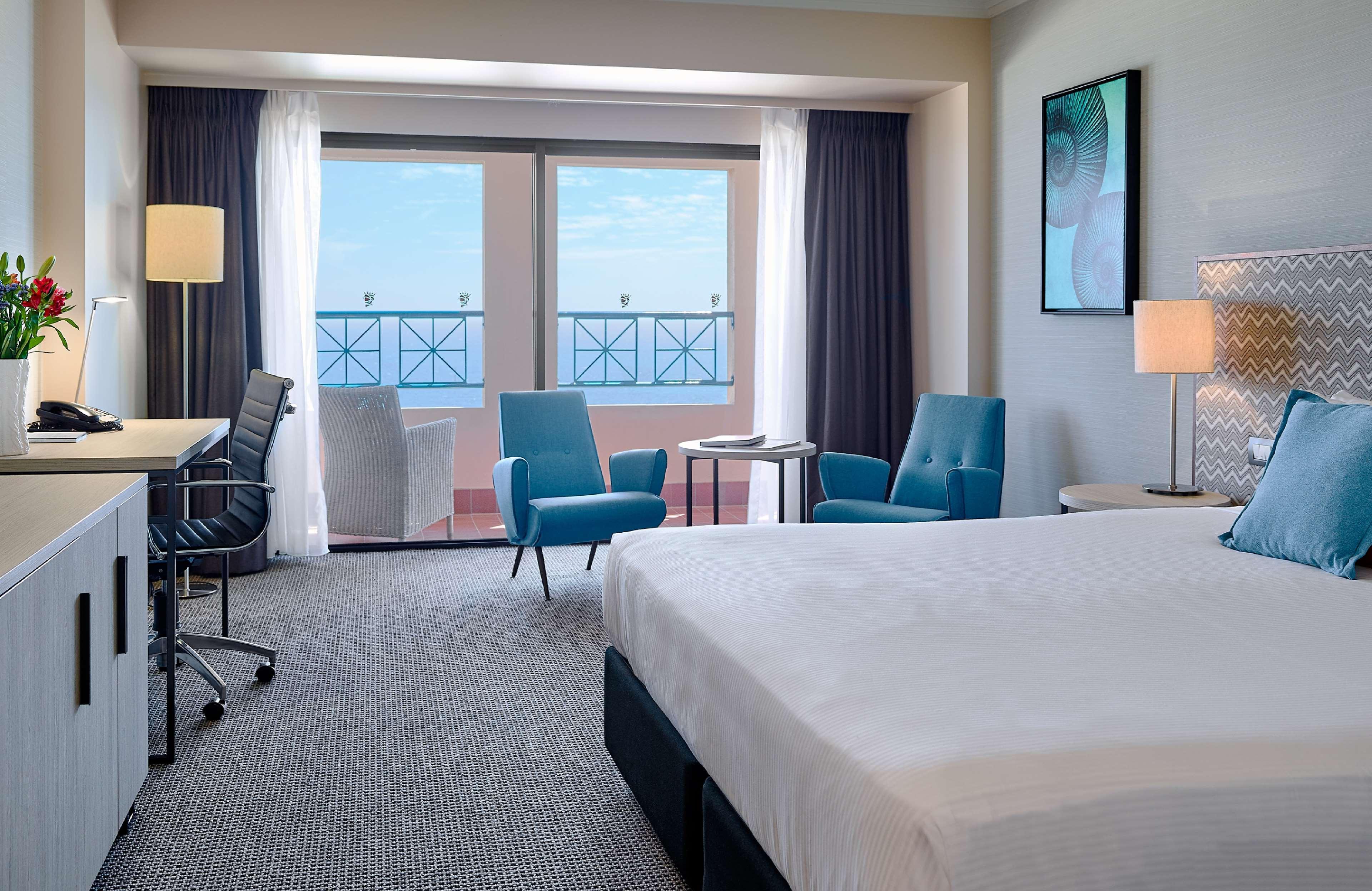 Stamford Grand Adelaide Hotel Glenelg Kültér fotó The image shows a hotel room featuring a large bed with a white duvet. There are two bright blue chairs and a small table near a large window that provides a view outside, possibly to a balcony. The room includes a workspace with a desk and an office
