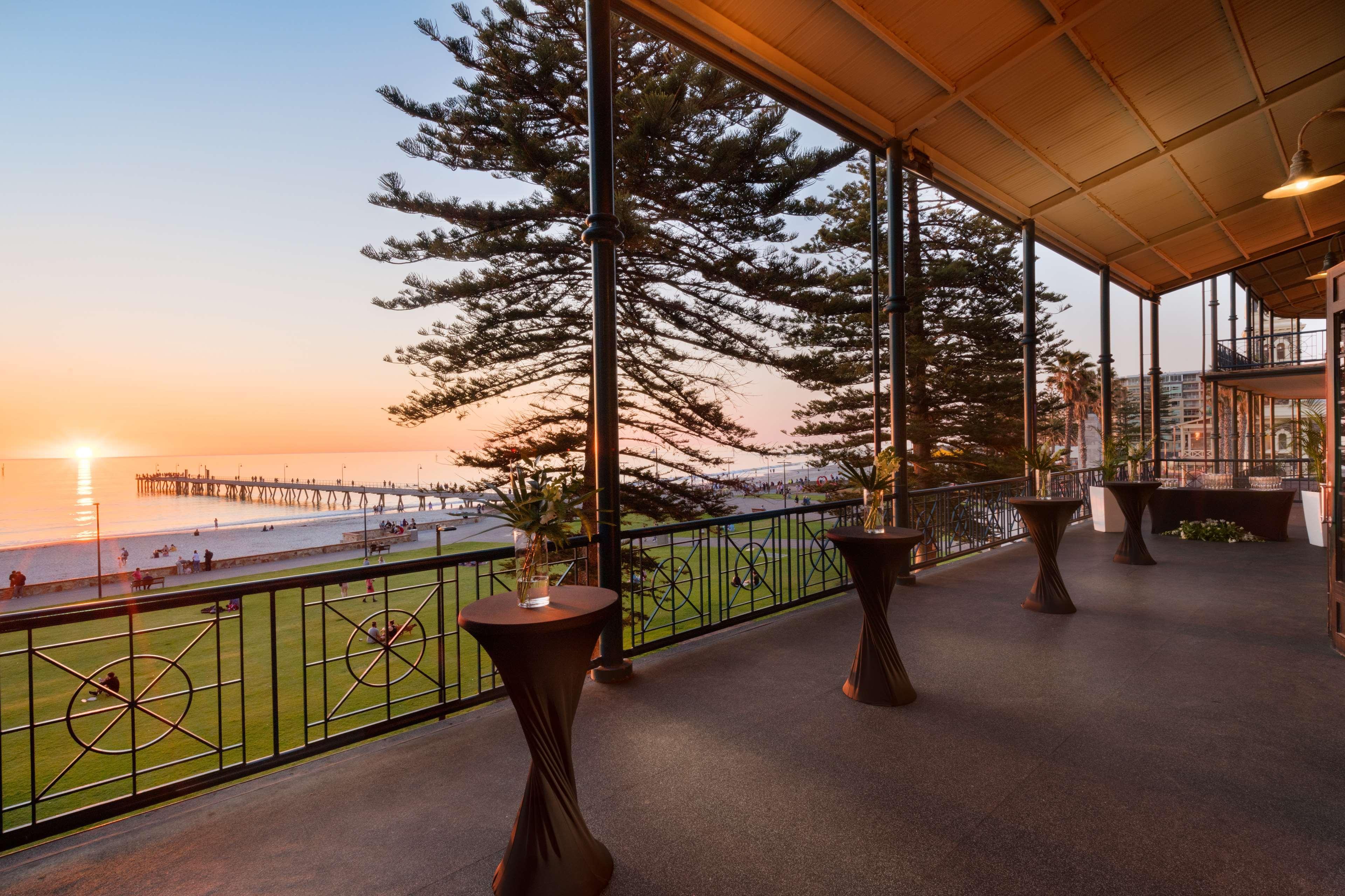 Stamford Grand Adelaide Hotel Glenelg Kültér fotó The photo shows a spacious indoor space with large windows overlooking a beach at sunset. The scene features a pier extending into the ocean and a horizon painted in warm colors from the setting sun. In the foreground, there are elegant tables with d