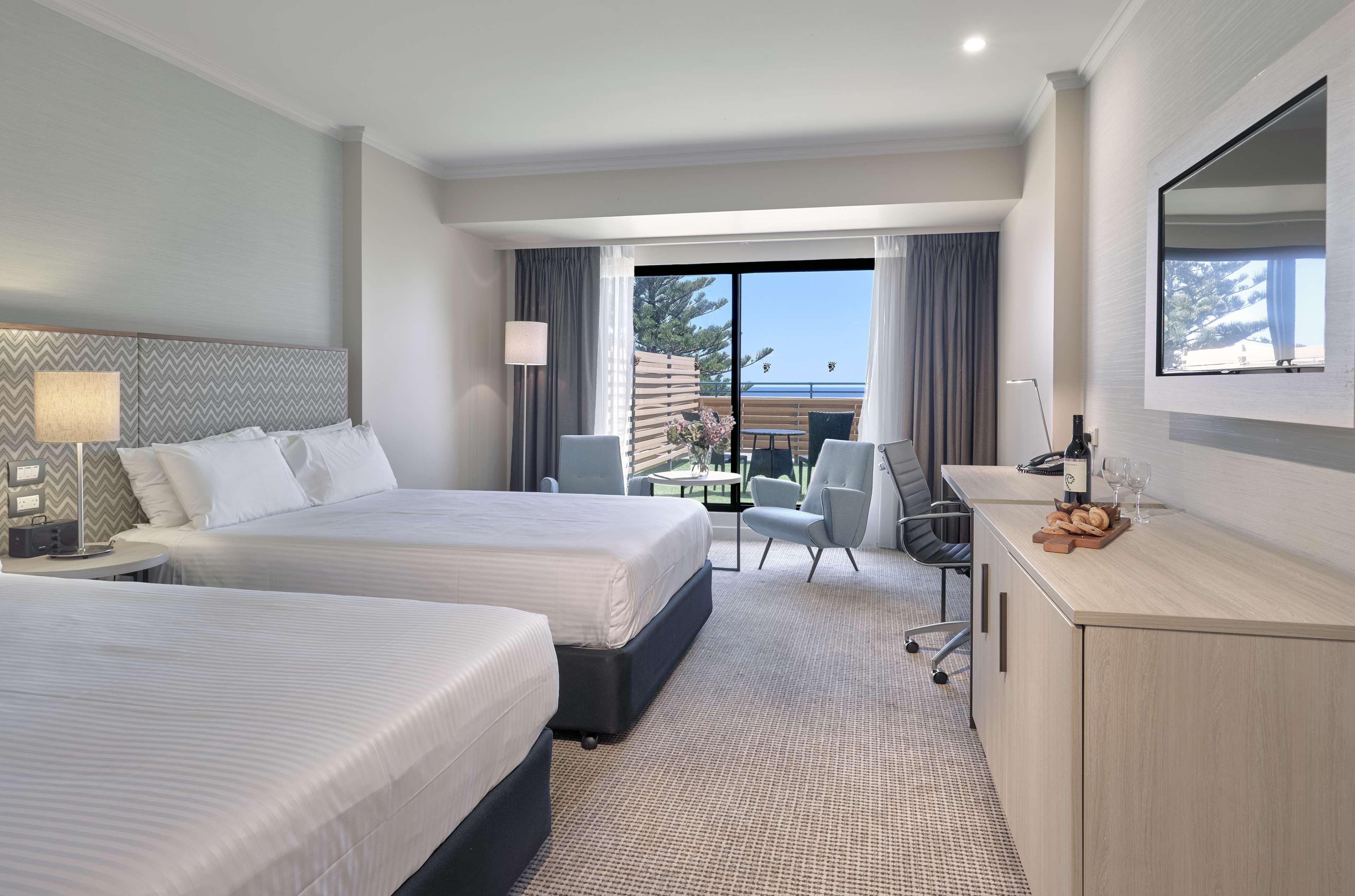 Stamford Grand Adelaide Hotel Glenelg Kültér fotó The photo shows a modern hotel room featuring two beds with white linens. There's a large window that provides natural light and a view of a balcony area. The room includes two chairs and a small table, creating a cozy sitting area. A television is m
