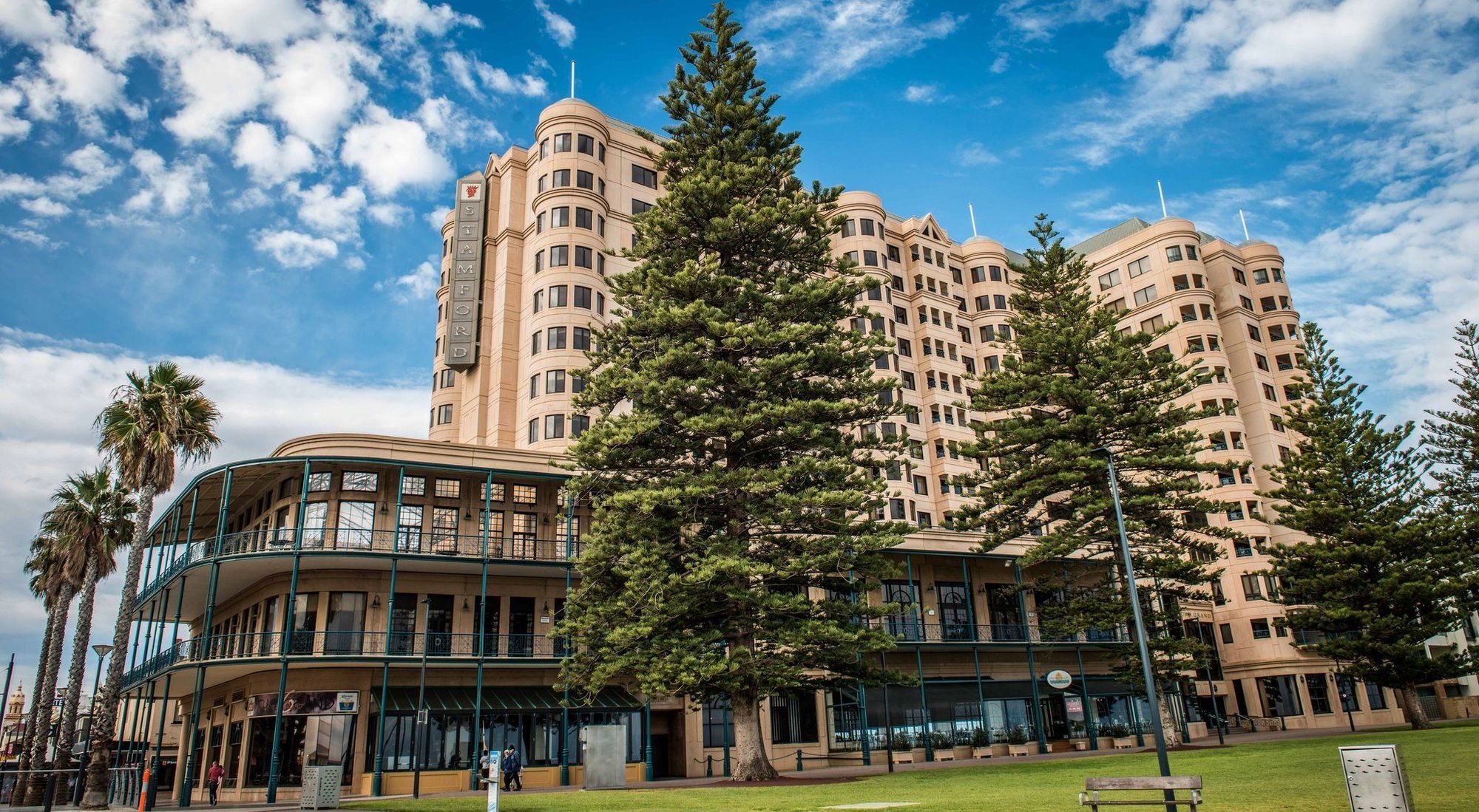 Stamford Grand Adelaide Hotel Glenelg Kültér fotó The image shows a large building with a distinctive architectural style, characterized by a tan or beige color. The structure appears to have multiple floors and rounded edges. In front of the building, there are several tall green trees, which add t