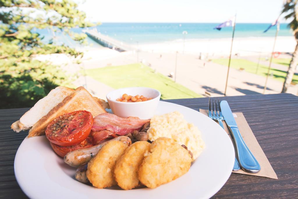 Stamford Grand Adelaide Hotel Glenelg Kültér fotó The photo shows a plate of breakfast food set against a beachside backdrop. The plate includes several items: crispy bacon, a grilled tomato, sausage links, golden hash browns, and a small serving of baked beans. There is also a piece of toast on the